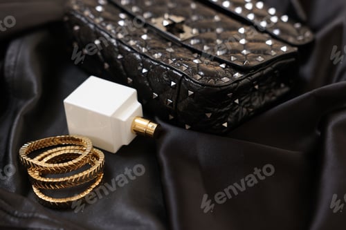 Preview: Leather bag, bottle of perfume and golden bracelets on black fabric, closeup. Space for text