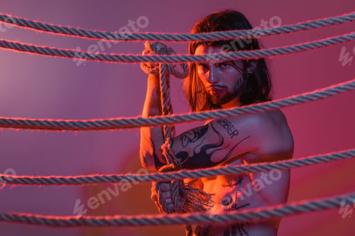Preview: Tattooed model holding rope and looking at camera on purple background with light