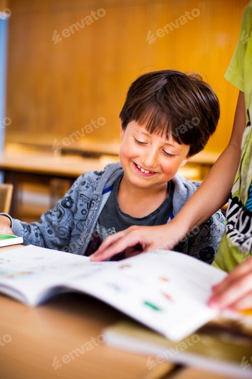 Preview: Smiling boy reading book while classmate standing at desk in classroom