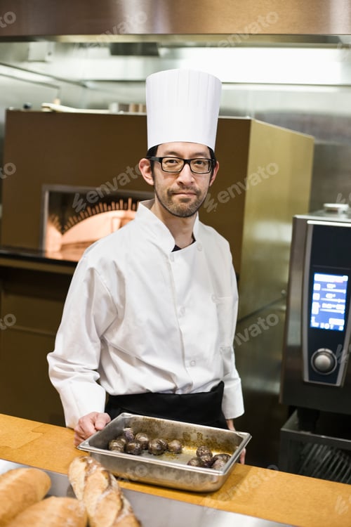 Preview: An asian chef working with a roasting pan of mushrooms in a commercial kitchen.