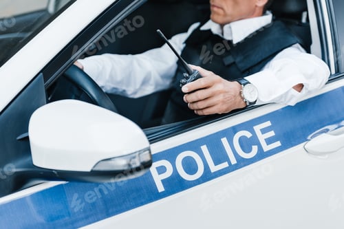 Preview: partial view of male police officer in bulletproof vest holding walkie-talkie in car