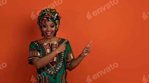 Preview: Pretty black woman in african costume pointing at copy space