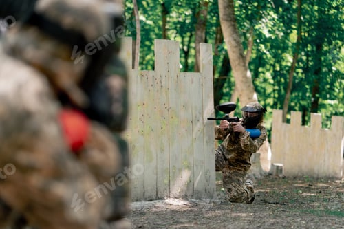 Preview: close up in the wild near tree structures player in a pintball uniform holds a pistol with a blue