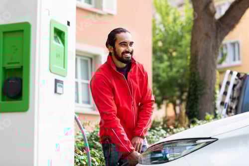 Preview: Smiling man in red jacket charging an electric car outdoors near a green station. Germany