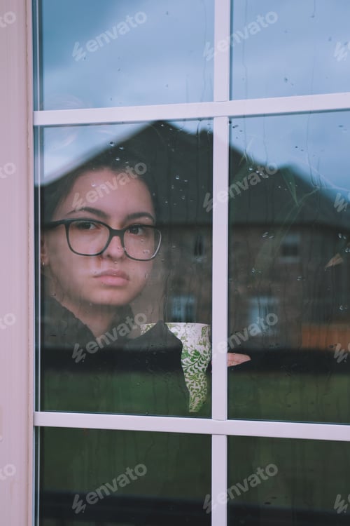 Preview: Young girl in glasses is holding a cup of tea or coffee and looking through a window on a rainy day