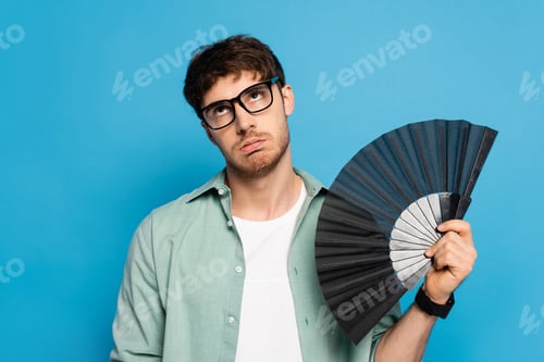 Preview: exhausted young man holding hand fan while suffering from summer heat on blue