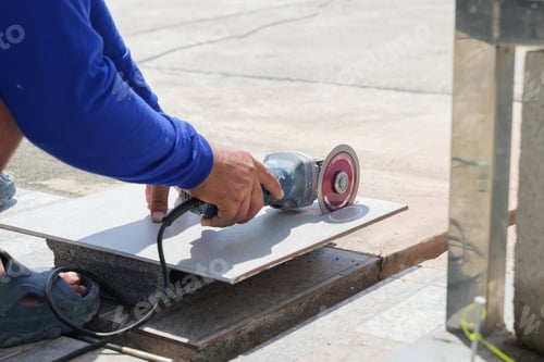 Preview: Worker in blue clothing uses an angle grinder to cut a gray tile on a piece of stone. Close-up of