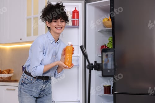 Preview: Smiling food blogger with bottle of juice recording video in kitchen