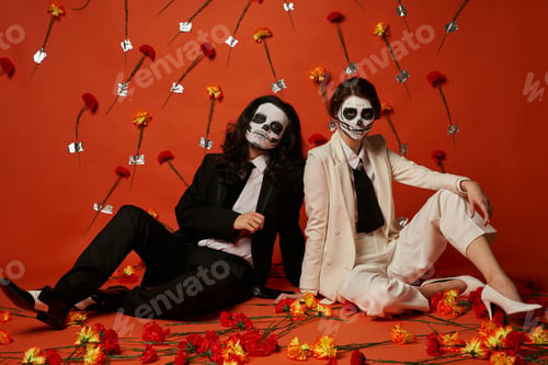 Preview: couple in sugar skull makeup and elegant suits sitting on floor in red studio with carnation flowers