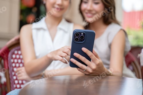 Preview: Blurred asian woman taking photos or showing phone to friend while sitting at table in front of cafe