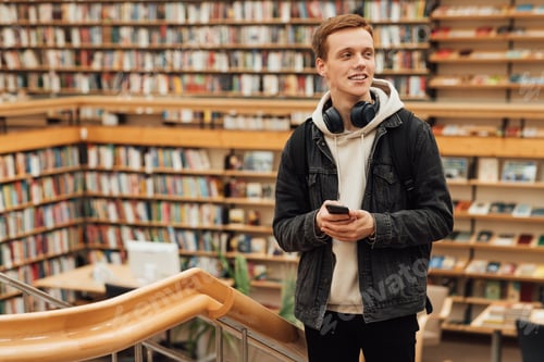 Preview: Smiling student with headphones and smartphone standing in university library