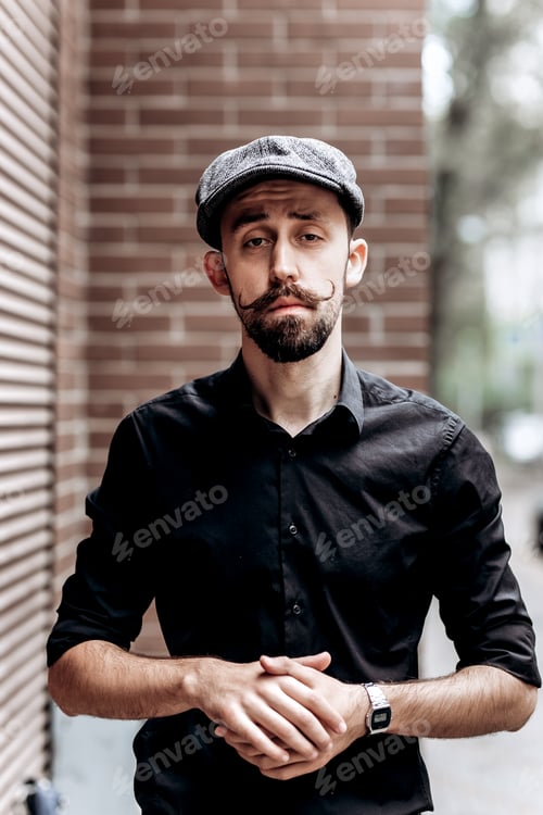 Preview: Stylish barber with mustache and beard dressed in a black shirt and flat cap stands on brick wall