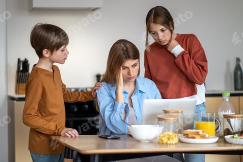 Preview: Mom with headache working on laptop on kitchen with children standing near looking at screen.
