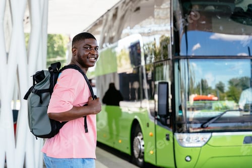 Preview: A young man wearing a pink shirt and a backpack stands next to a green bus