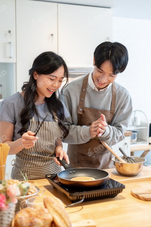 Preview: A joyful Asian couple is having fun while making pancakes in the kitchen together.