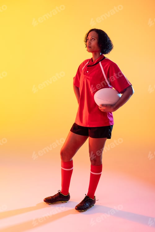 Preview: African american female rugby player with rugby ball over neon yellow lighting