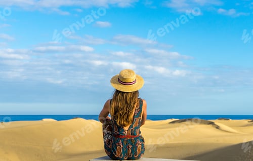 Preview: Horizontal shot of young female with a straw hat standing on a sandy beach under a clear bright sky