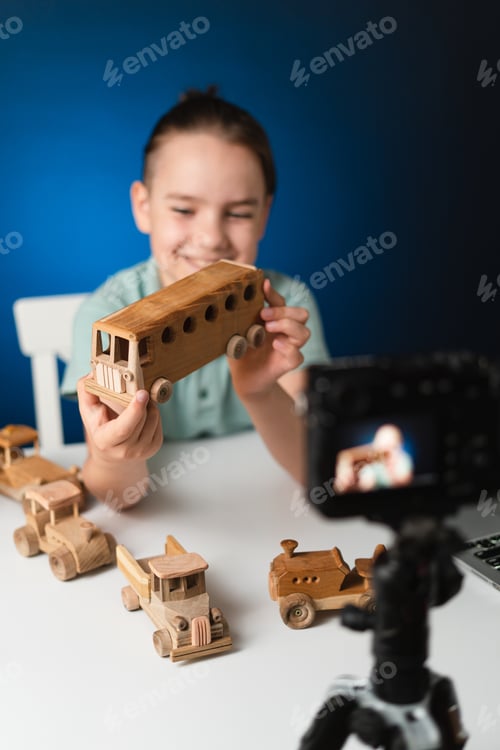 Preview: Kid blogger filming video on camera indoors. Promoting eco wooden toys. Boy holding a bus in hands.