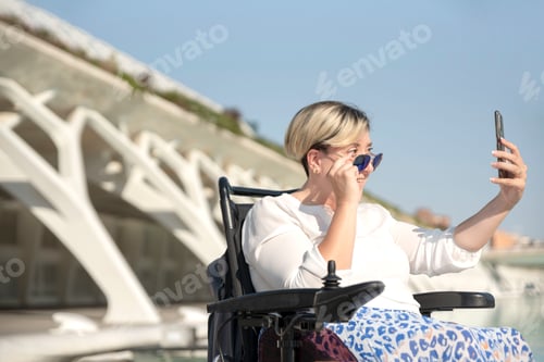 Preview: a smiling woman with a disability in a wheelchair taking a selfie looking behind sunglasses