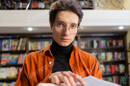 Preview: portrait of a young guy in glasses with a book in his hands in the library preparing exams