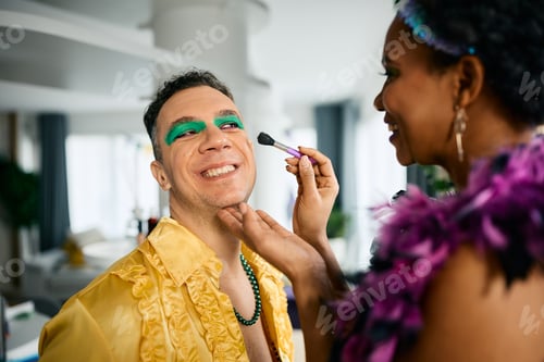 Preview: Happy man having fun while his friend is putting him Mardi Gras make-up at home.