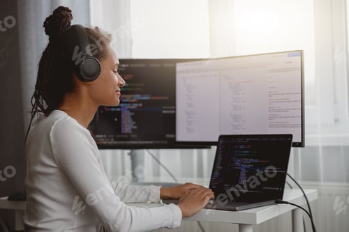 Preview: Cheerful beautiful female IT developer listening to music in headphones while coding at home