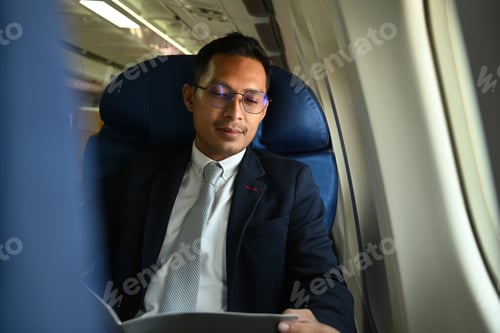 Preview: Asian businessman in formal suit sitting near window in an airplane and reading financial document.
