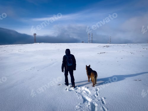 Preview: Rear view of man with backpack hiking in the mountains with his German shepherd dog during winter