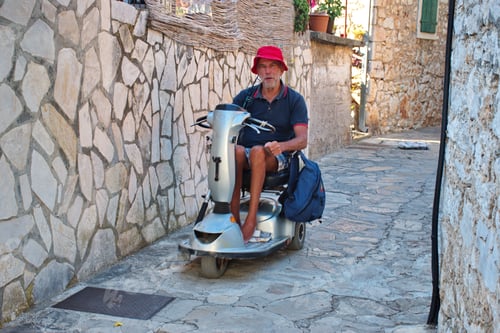 Preview: Portrait of senior man in electric wheelchair driving through the narrow Mediterranean street