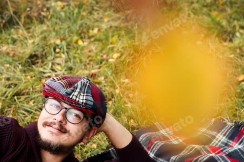 Preview: Young man in autumn wearing cap. Man lying in nature during autumn. Blurred yellow fall background.