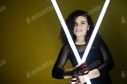 Preview: Curly brunette plus size model with two long led lamp against yellow wall.