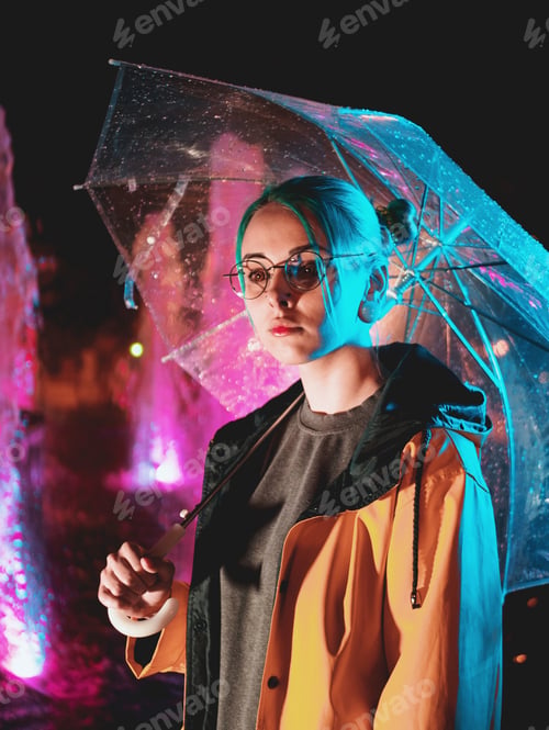 Preview: girl with blue dyed hair in raincoat and with transparent umbrella. Night neon illumination of city