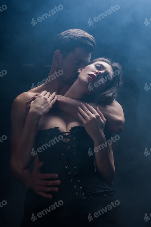 Preview: Handsome man kissing and hugging seductive woman in corset on black background with smoke