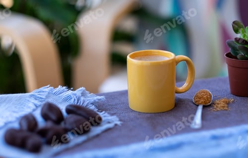 Preview: Hot coffee in yellow ceramic cup, outdoor on balcony with plants and chairs - defocused chocolates