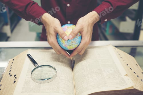 Preview: Young student man reading book and holding earth model in hand.