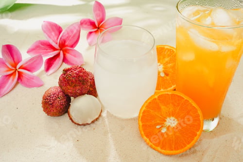 Preview: Closeup shot of a glass of orange juice and a cocktail next to lychee, oranges and plumeria flowers