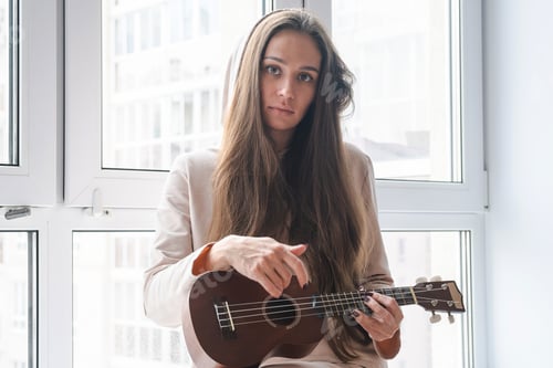Preview: Woman with long hair plays ukulele by window. Concept of music enjoyment.