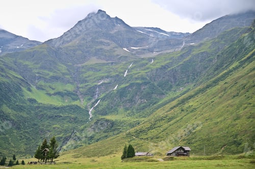 Preview: Wooden lodge with a high mountain and waterfall behind, lush green grass in Austrian Alps