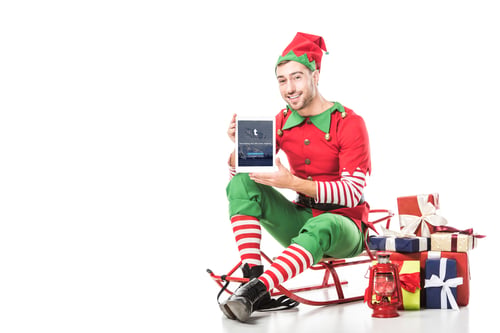 Preview: man in christmas elf costume sitting on sleigh and holding tablet with tumblr app on screen isolated