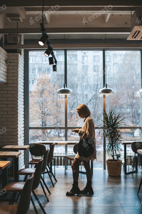 Preview: Young blonde woman texting message using smartphone in modern cafe or co-working place.