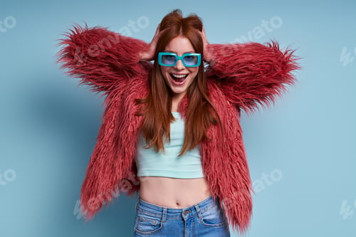 Preview: Happy young woman in fluffy coat holding hands in hair against blue background