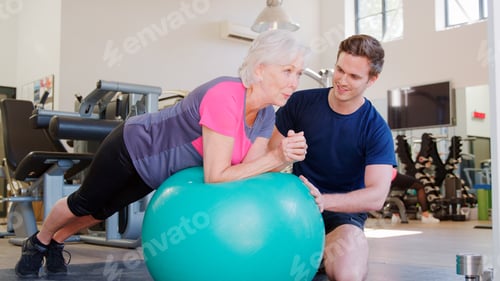 Preview: Senior Woman Exercising On Swiss Ball Being Encouraged By Personal Trainer In Gym