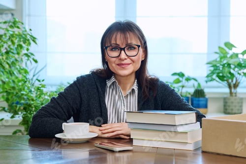 Preview: Middle aged woman holding cup of coffee, sitting at table with pile of books