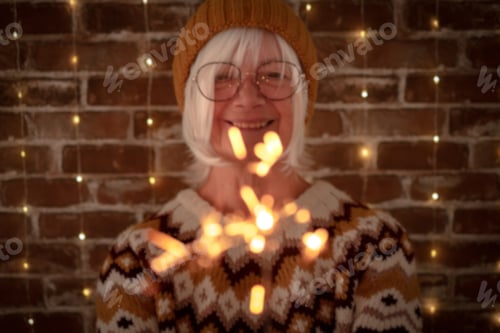 Preview: Blurred senior caucasian woman with eyeglasses and winter sweater holding sparkler light