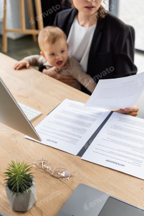 Preview: blurred businesswoman holding document while sitting at work desk with toddler daughter