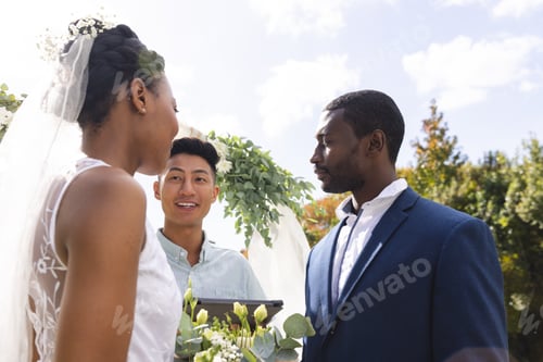 Preview: Happy diverse male officiant with bride and groom at outdoor wedding ceremony in sunny garden