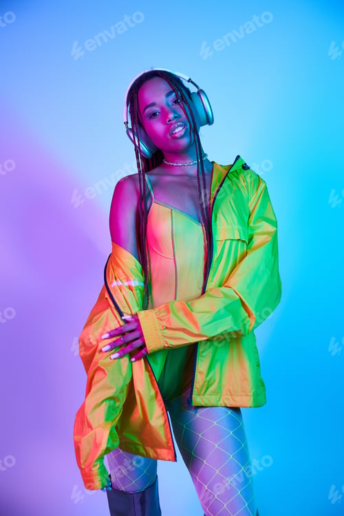 Preview: charming dark-skinned woman in wireless headphones posing in jacket in studio with neon lights