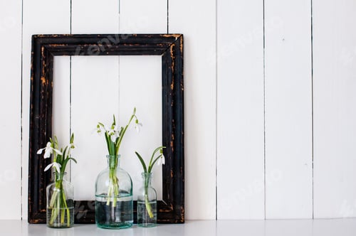 Preview: White Spring Flowers Snowdrops In Vintage Glass Bottles And A Blck Frame On An White Barn Wall