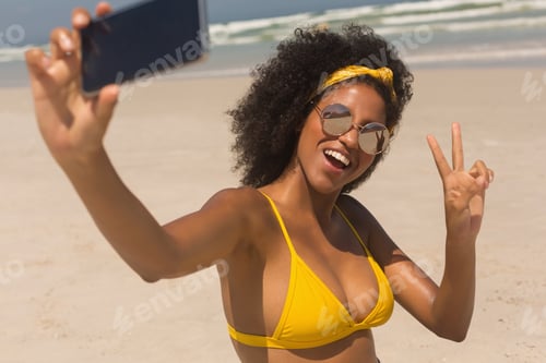 Preview: Young African American woman in yellow bikini and sunglasses taking selfie with her mobile phone