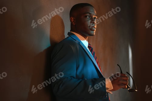 Preview: Retro African American Businessman In Blue Suit Holding Sunglasses. Leaning Against Gray Wall.